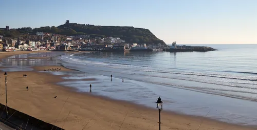 Scarborough beach morning This landscape photograph captures Scarborough beach in Yorkshire during a calm spring morning. The soft sunlight illuminates the sandy shoreline, creating gentle reflections on the wet sand as small waves lap at the edge. In the background, Scarborough Castle is prominently perched atop the hill, overlooking the seaside town and adding a historical element to the scene. The town's buildings cluster along the waterfront, leading towards the harbour where the gentle sea meets the sky. The image showcases both the natural beauty of the beach and the iconic castle, perfectly encapsulating the coastal charm of Scarborough in Yorkshire.